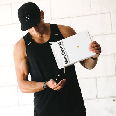 Man in black sleeveless top and shorts holding a white cereal box against a white brick wall.